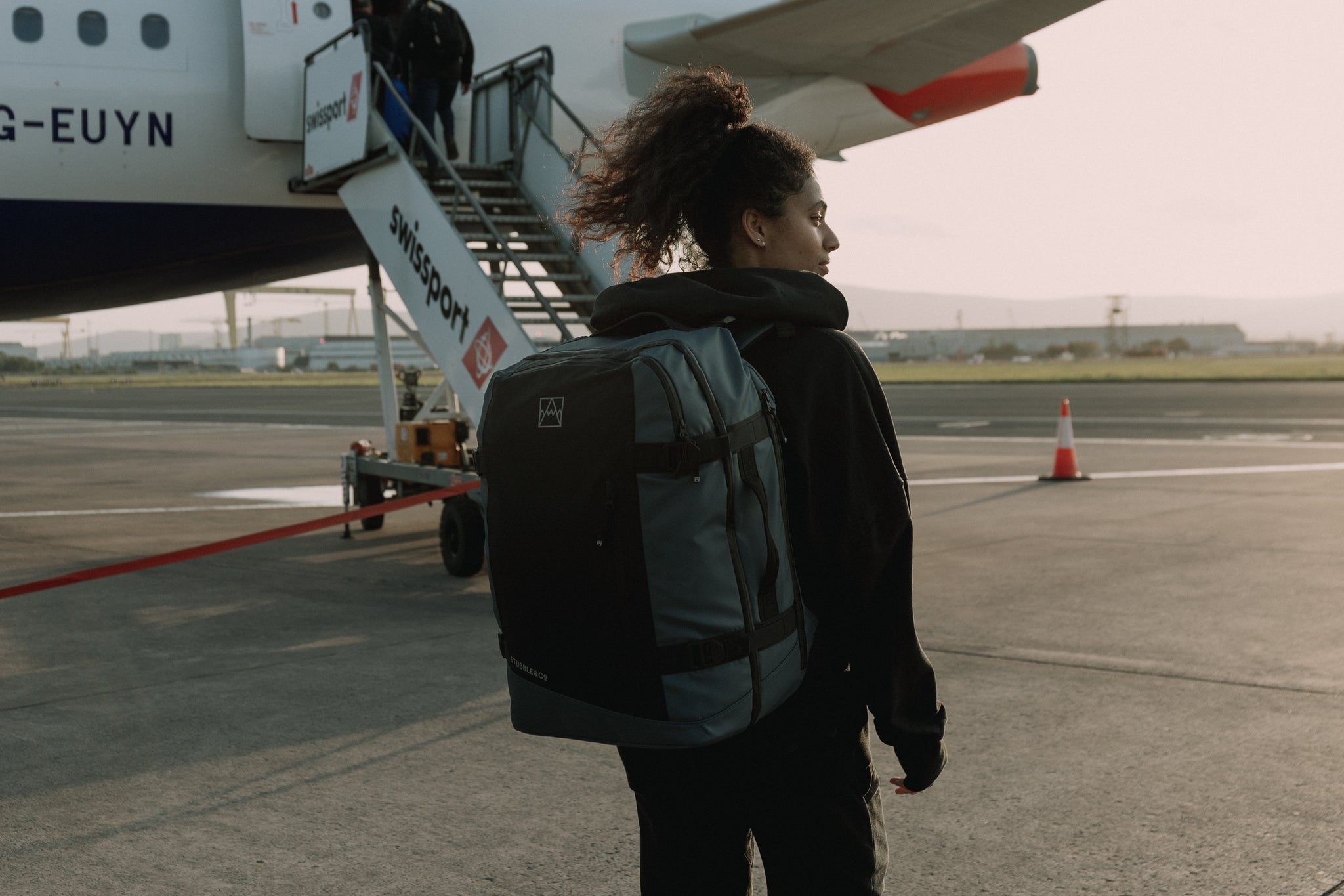 A woman standing in front of an airplane about to board wearing a tasmin blue adventure bag on her back