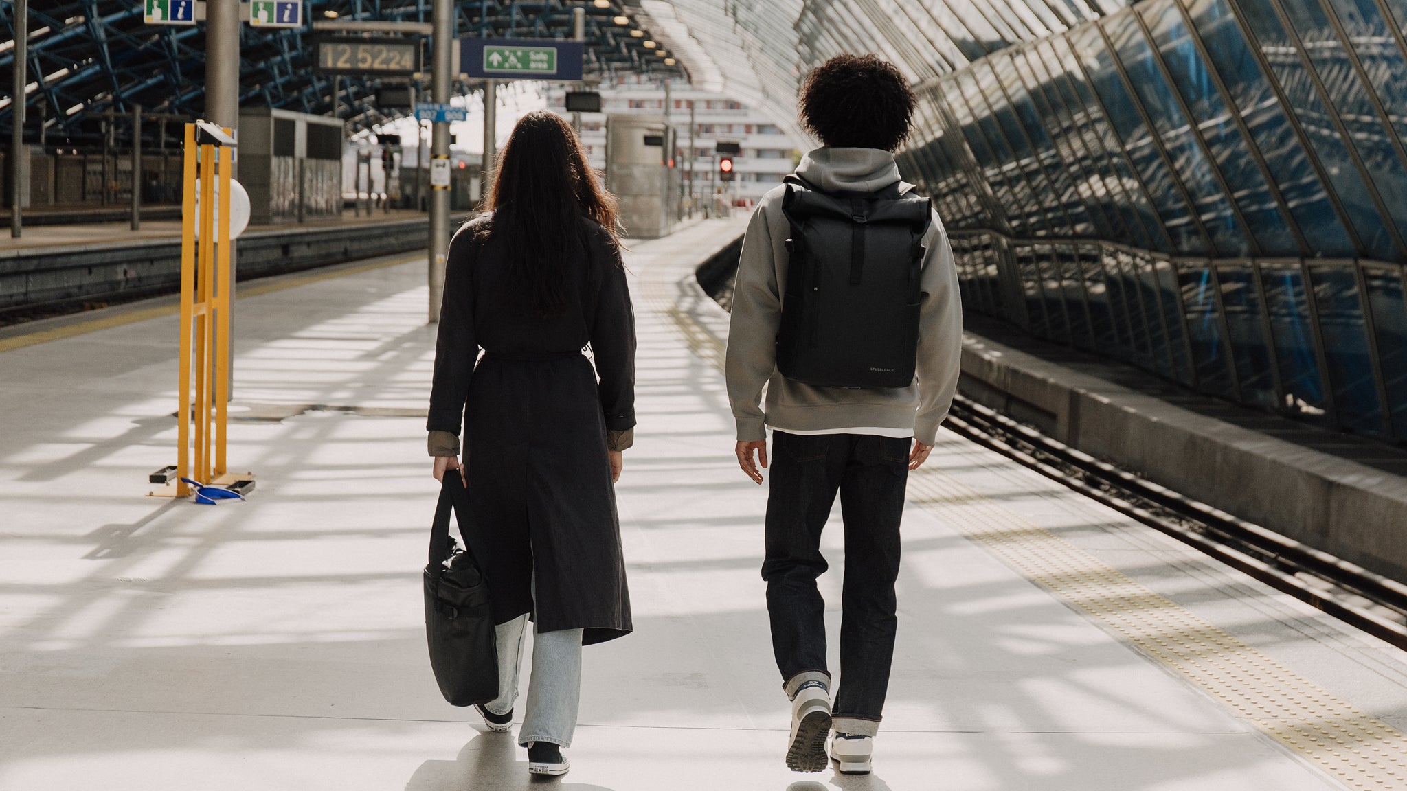 Man and women at a train station with a black backpack and tote bag