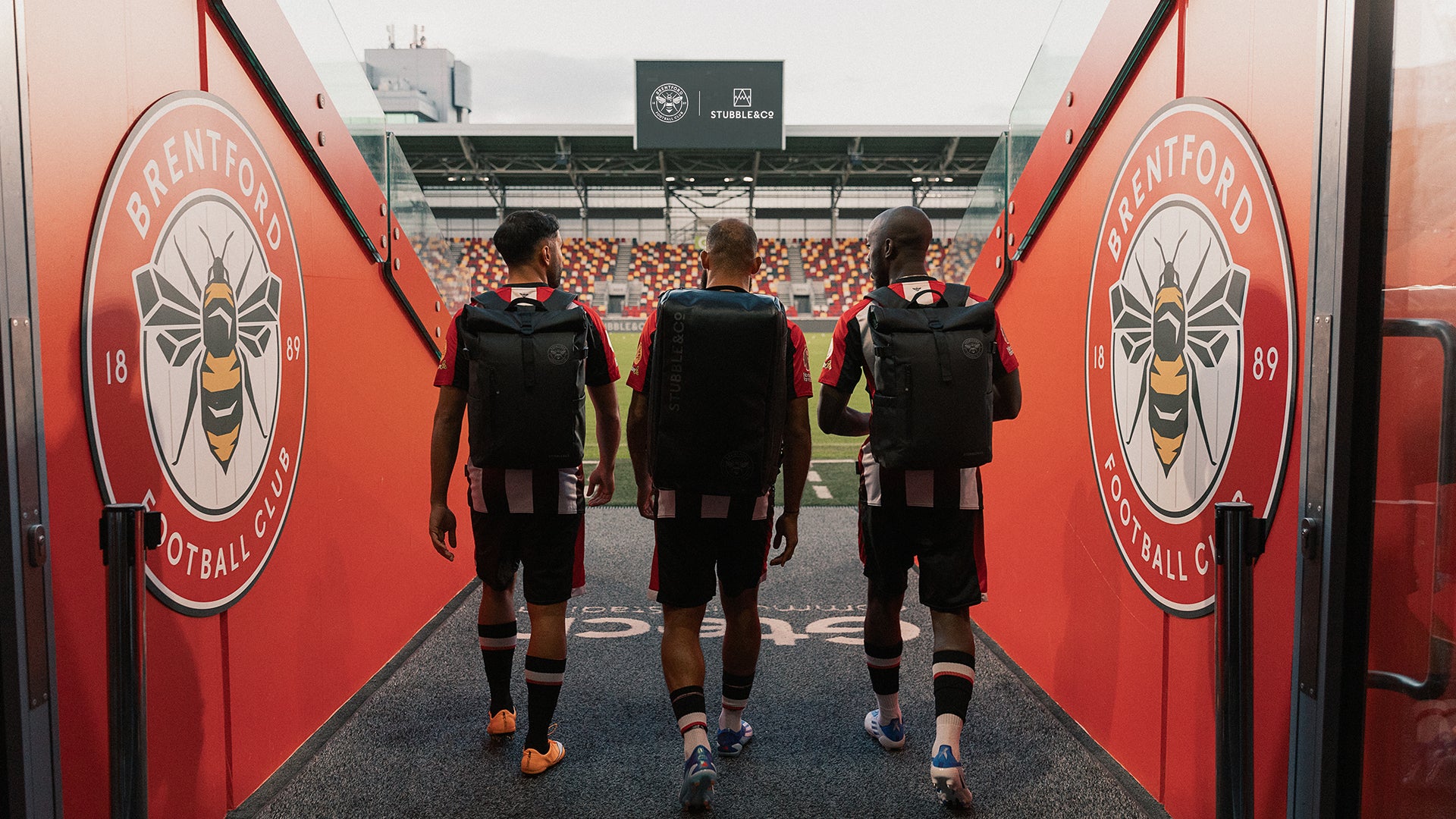 3 football players walking out towards the football pitch wearing black backpacks and rucksacks
