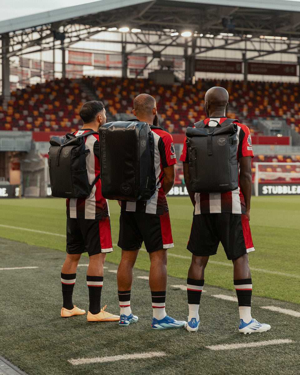 three brentford players facing away from the camera on the pitch wearing black rucksacks and backpacks