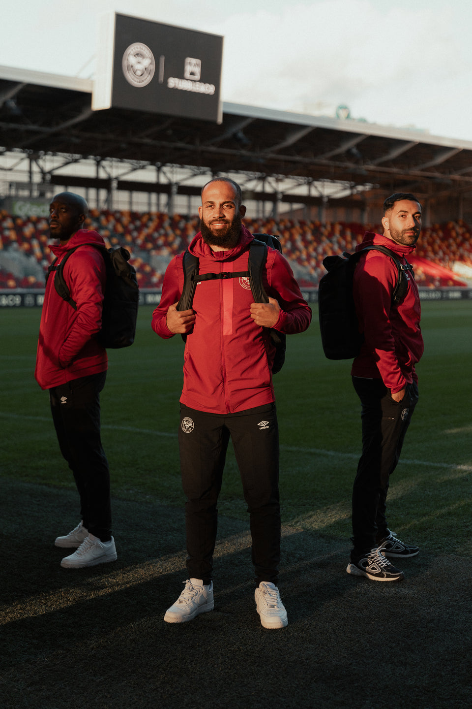 Three Brentford football players standing on the pitch wearing black backpacks and rucksacks