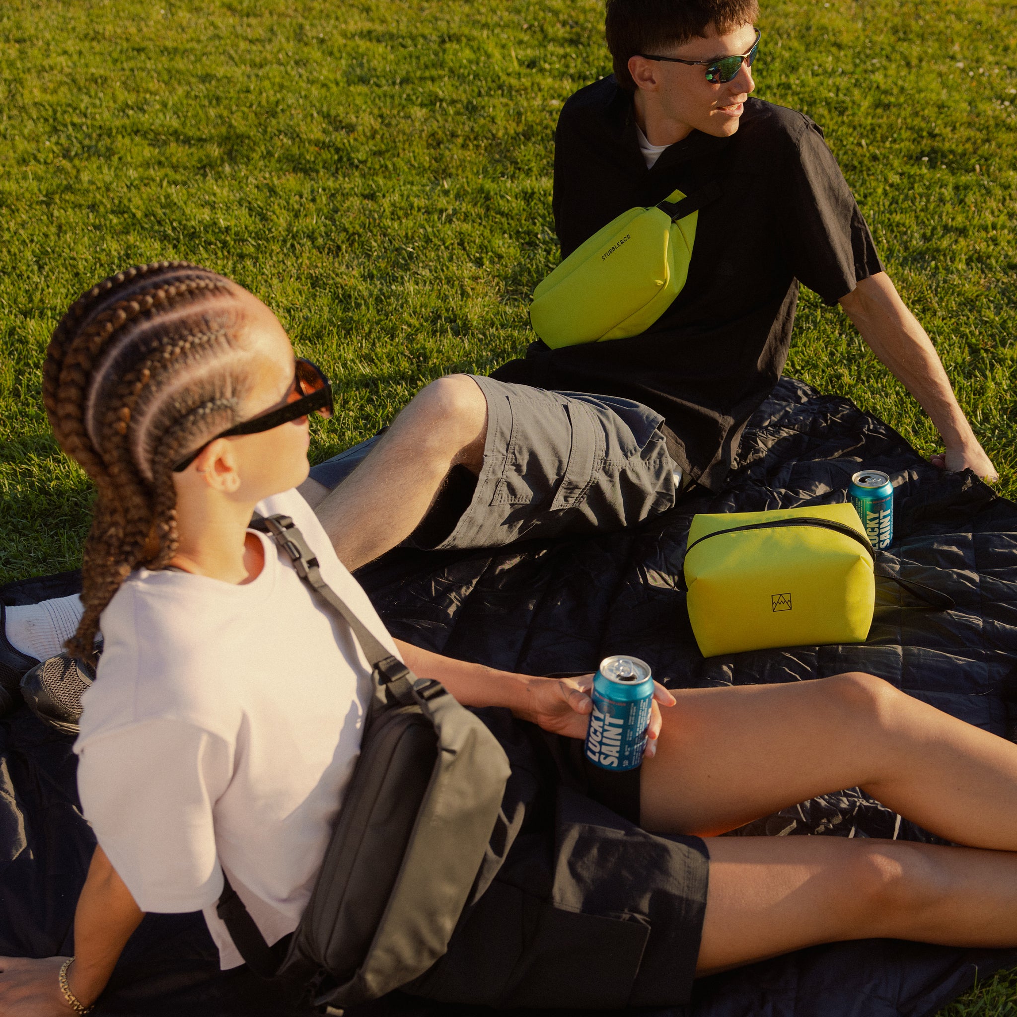 A man and woman having a picnic on grass wearing crossbody in Volt and with a Beer Bag next to them