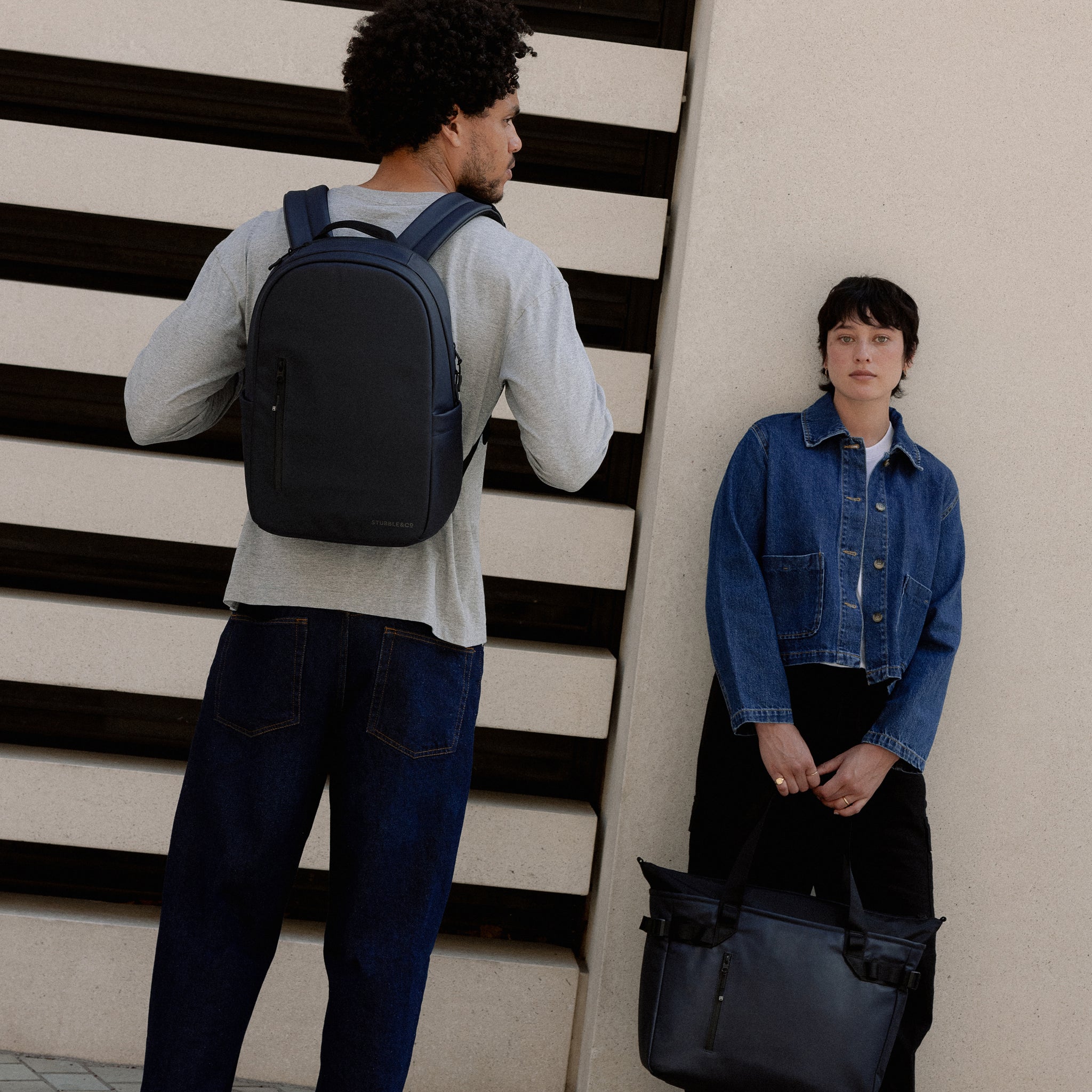 A man wearing an Everyday Backpack 15L in Midnight Blue on his back standing next to a woman holding a Laptop Tote Bag