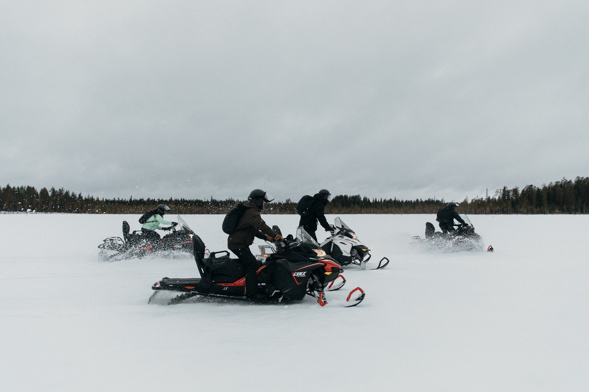 A group of 4 people driving on snowmobiles in the snow wearing black backpacks and rucksacks