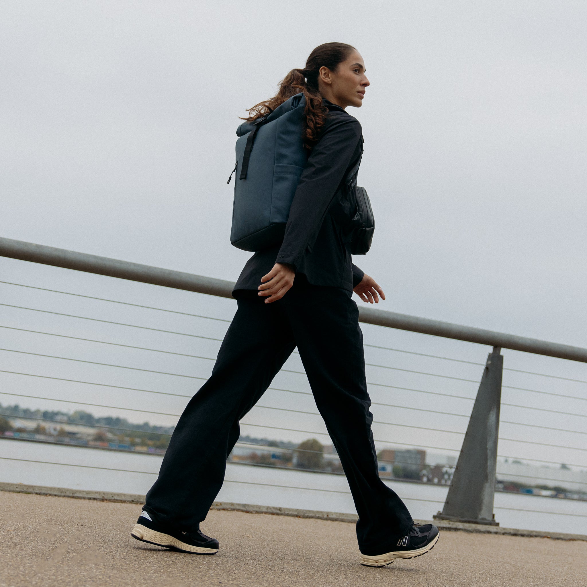 A woman walking next to a river wearing a Roll Top 15L backpack in Tasmin Blue on her back