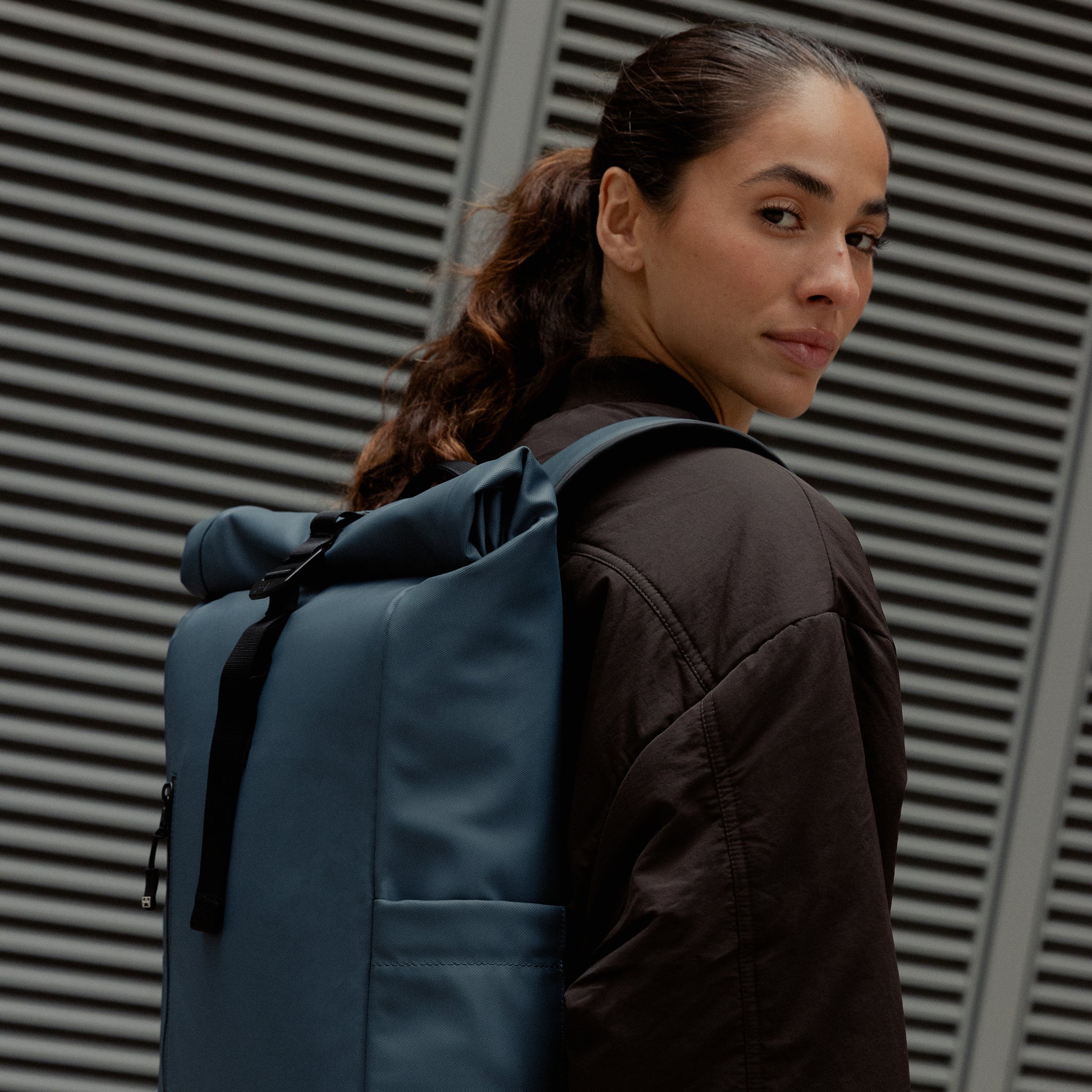 A woman standing side on wearing a Roll Top 15L backpack on her back in Tasmin Blue
