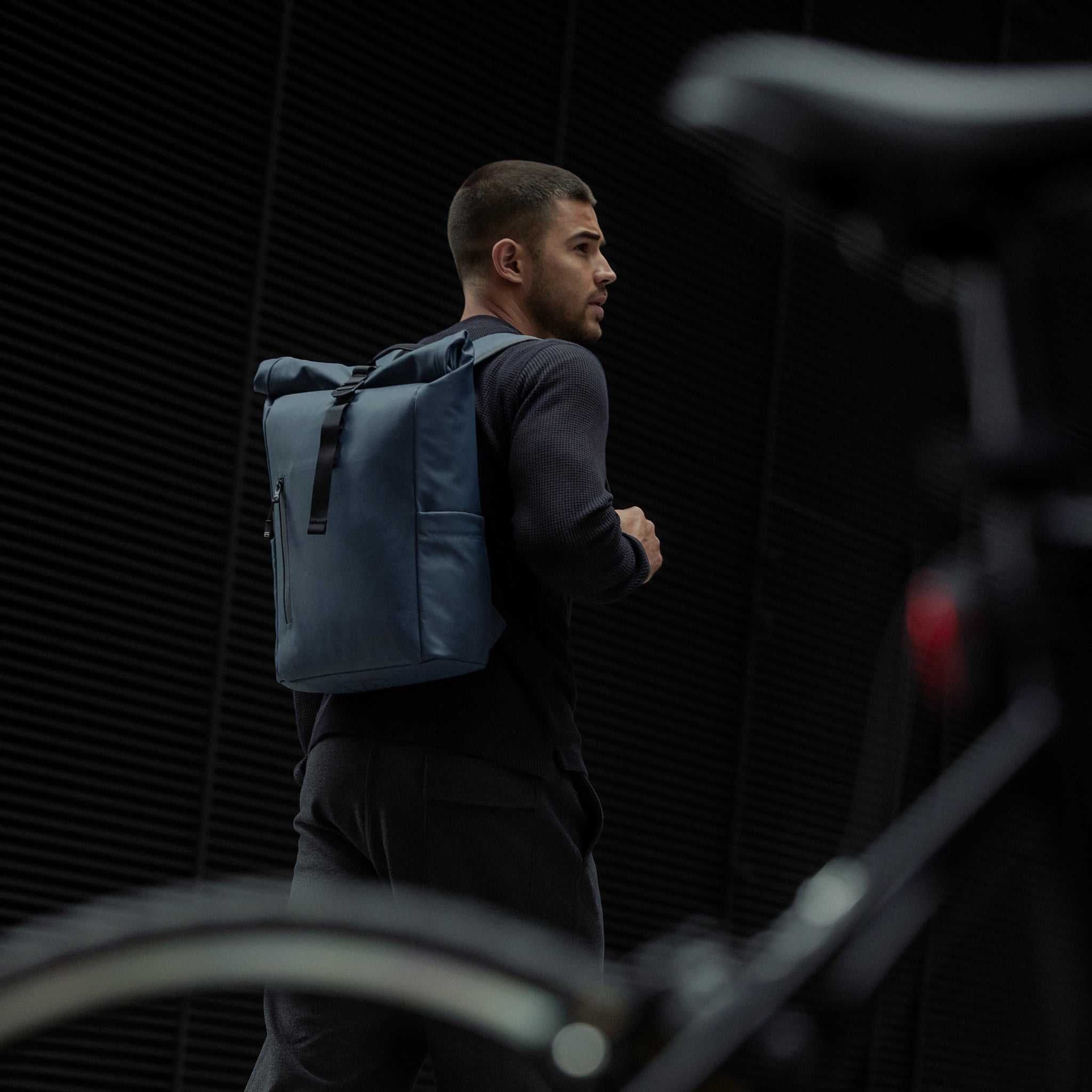 A man standing next to a bike with a Roll Top 15L backpack in Tasmin Blue on his back