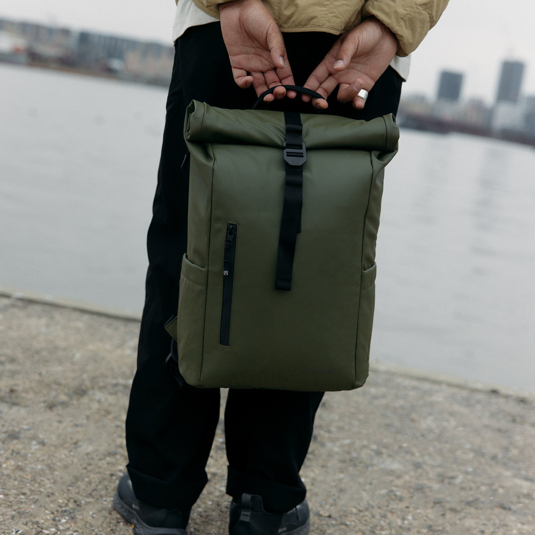 A man holding a Roll Top 15L backpack in Urban Green by his side