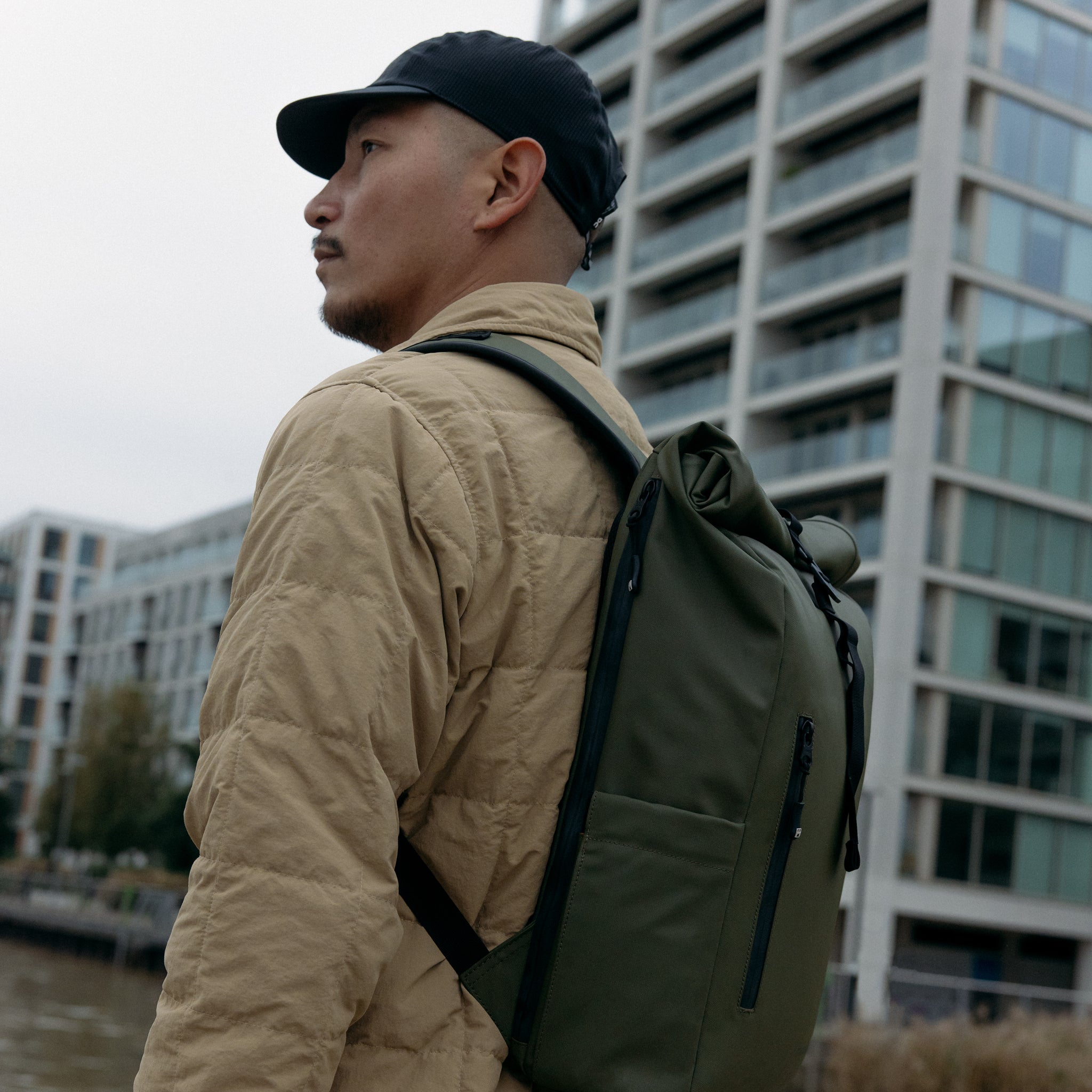 A man standing side on with a Roll Top 15L backpack on his back in Urban Green