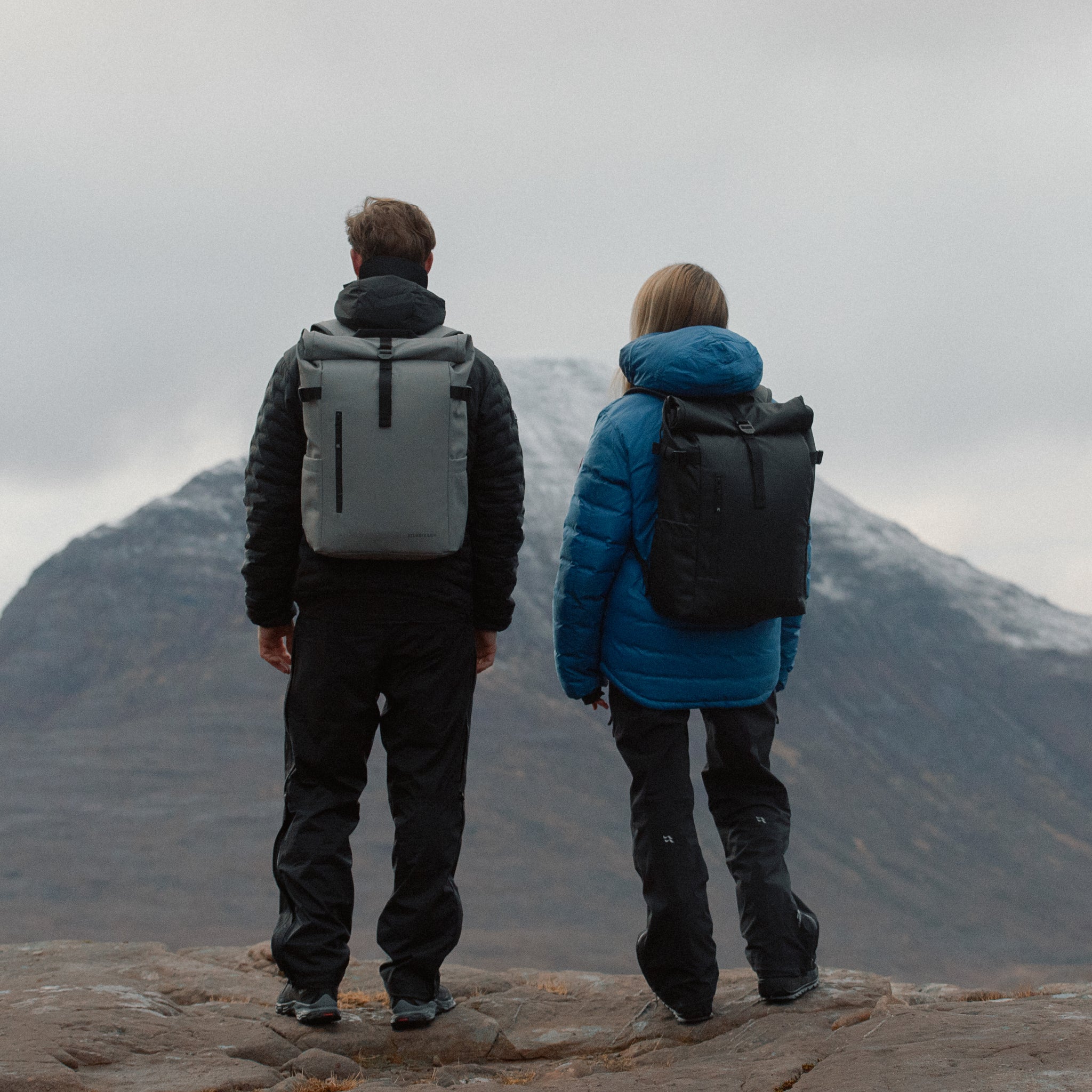 Man and women wearing The Roll Top 20L Backpack in All Black on a mountain