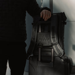 Person putting The Roll Top 20L Backpack in All Black on a suitcase using a trolley sleeve