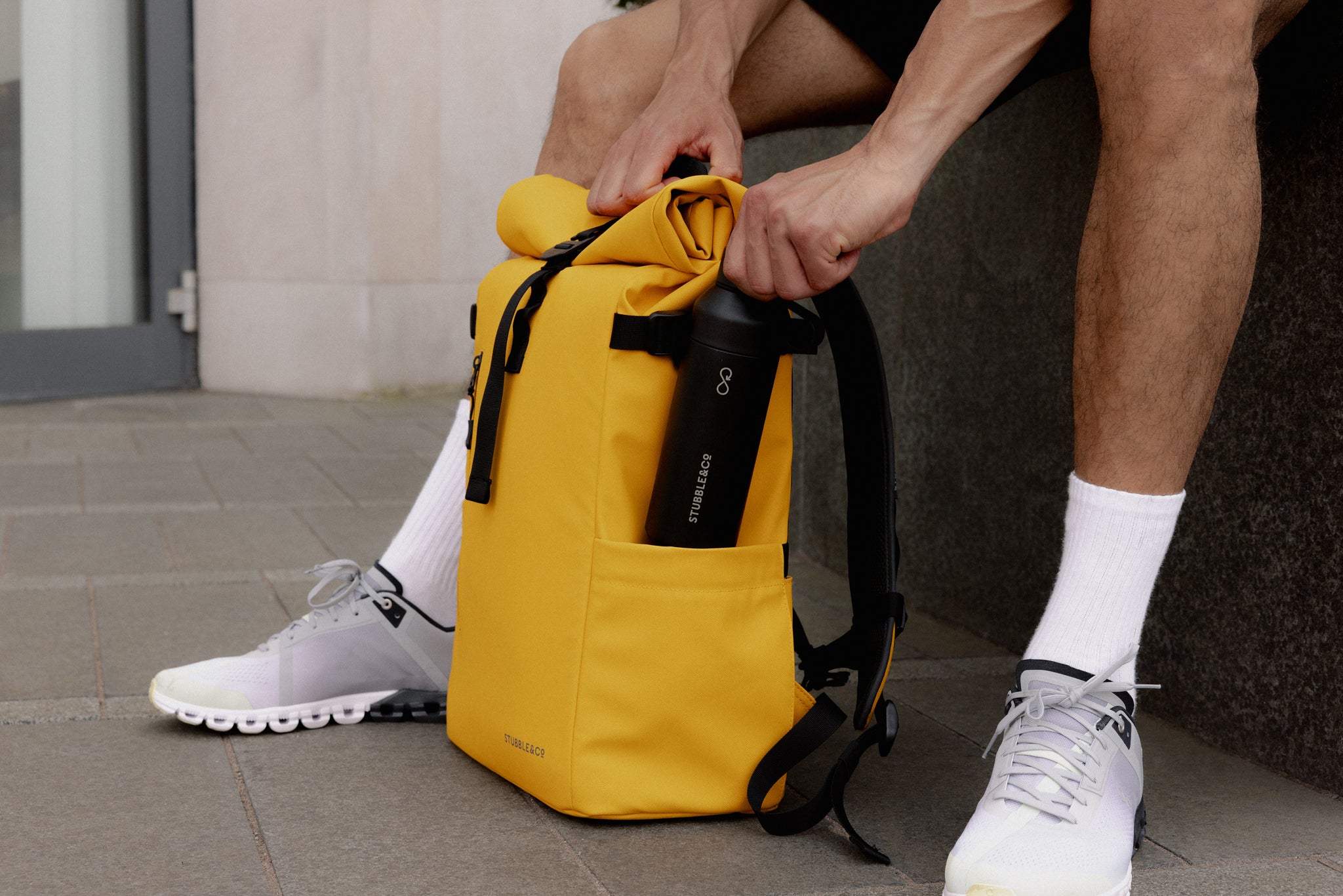 A man putting a stubble and co water bottle into the elasticated pocket on the side of a yellow roll top 30l