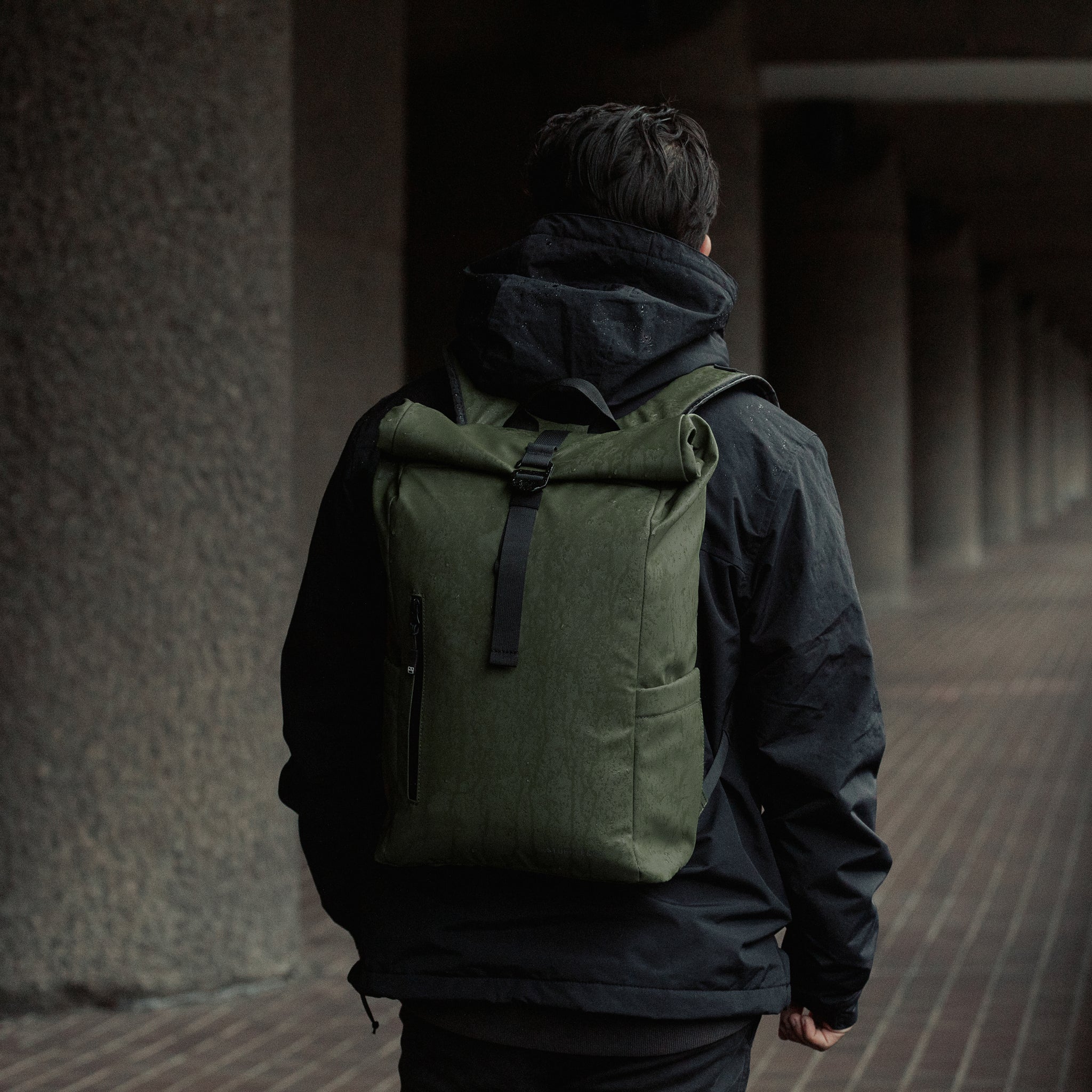 A man walking through the rain with a Roll Top 15L backpack in Urban Green on his back