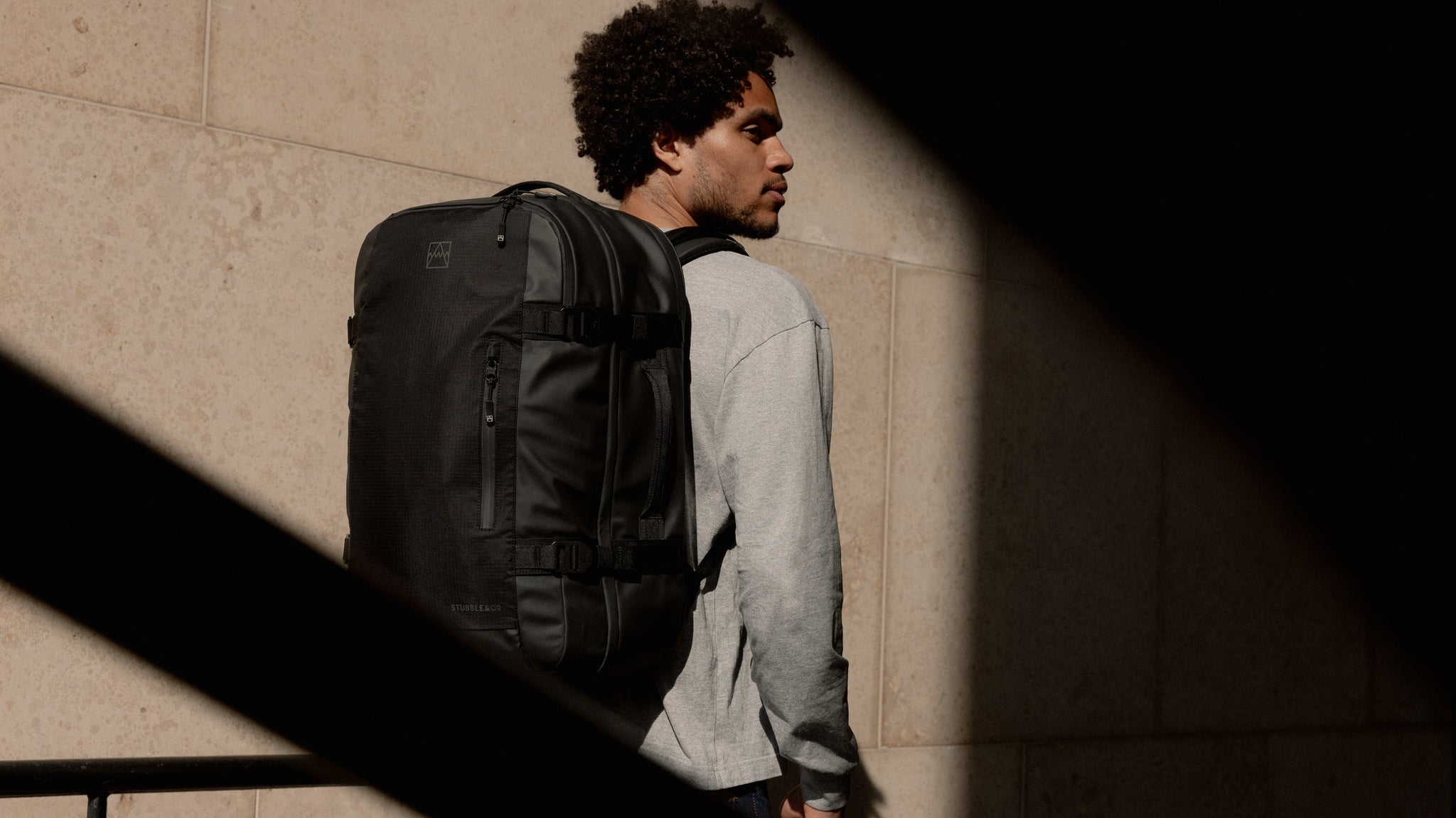 A man walking down the street in the sunshine wearing a Black travel Backpack 40L on his back