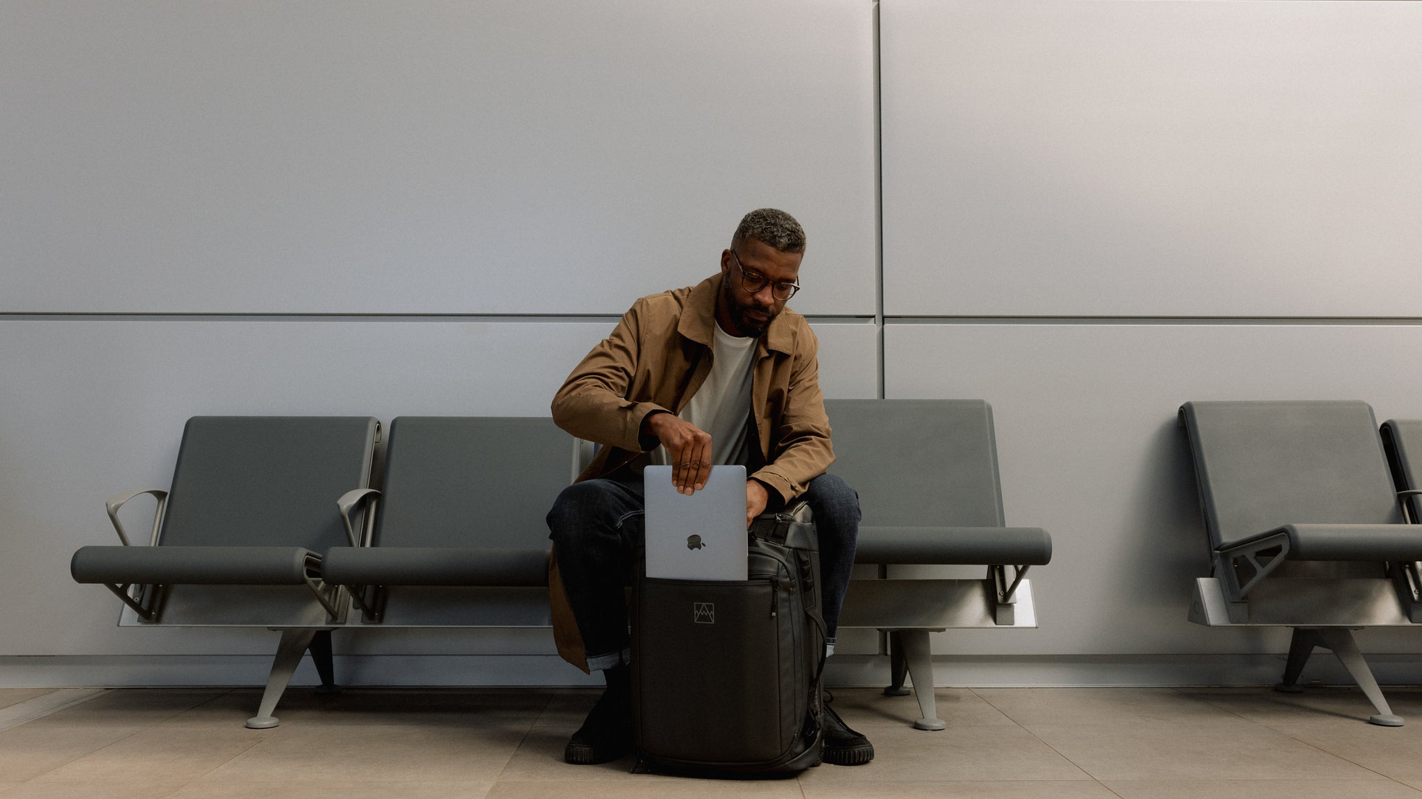A man sitting in an airport terminal putting his laptop into the front of a Wheeled Kit Bag 40L