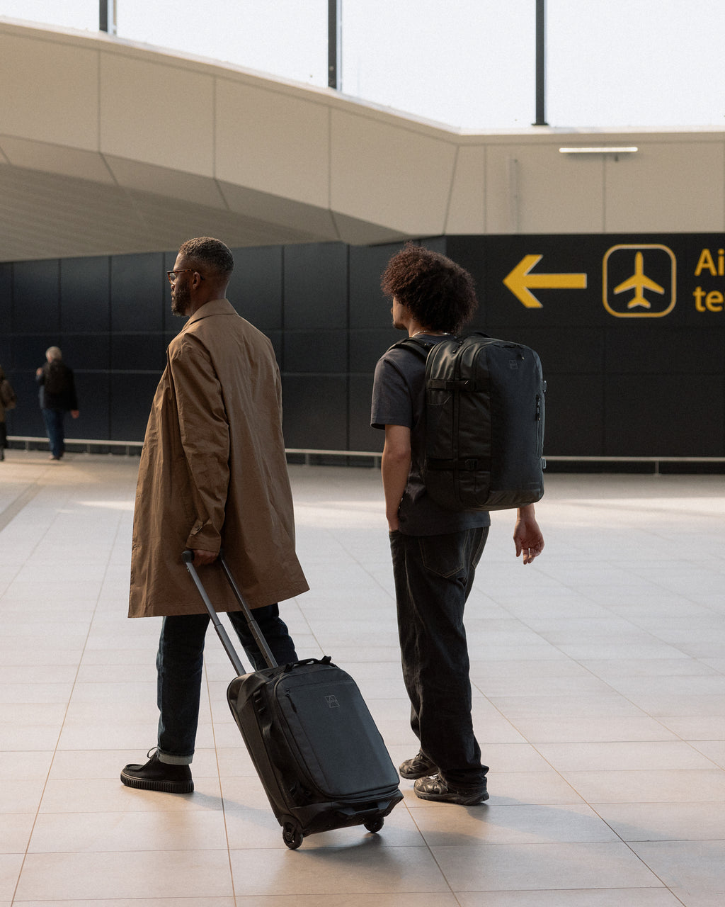 a man wheeling an Wheeled Kit Bag 40L in All Black and another man wearing a Travel Backpack 40L on his back