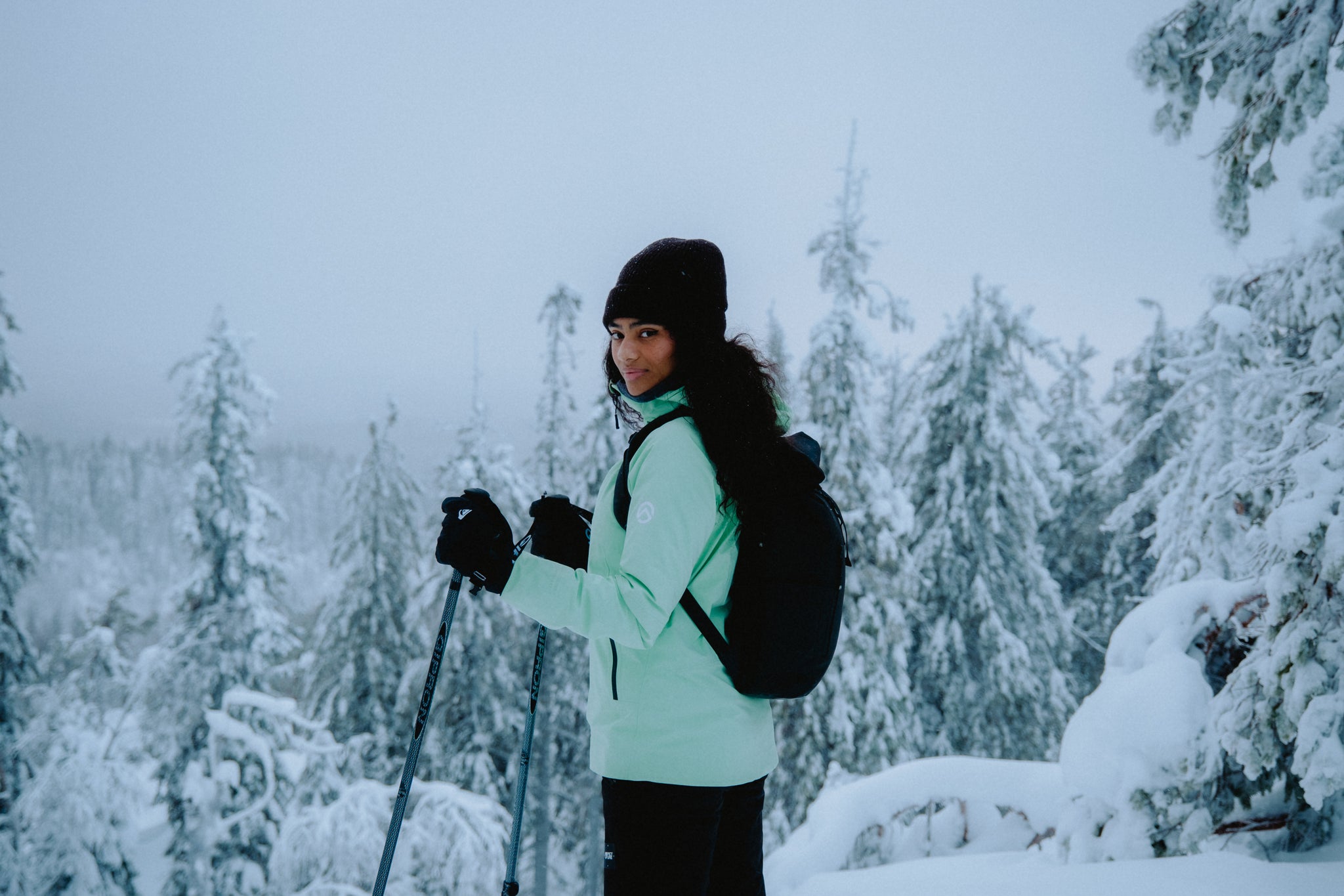 A women standing with snow and trees surrounding her whilst wearing a black backpack