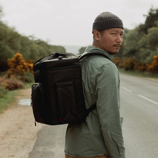 Man wearing cooler backpack in black walking by a road
