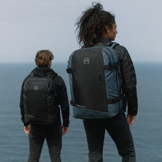 Shot from the back, a female model in the foreground wearing the Tasmin Blue Adventure Bag, with a man in the background wearing the All Black Adventure Bag.