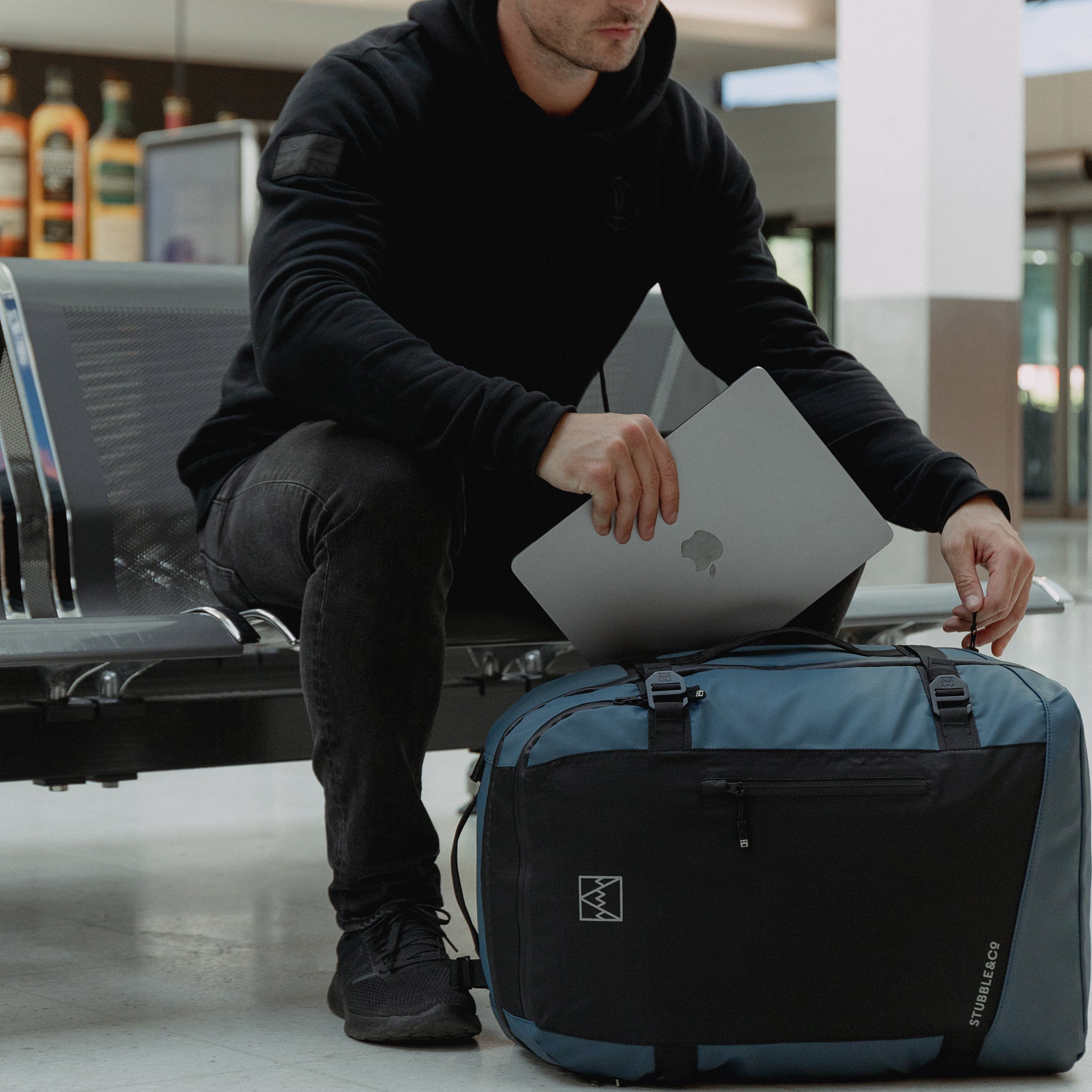 A man sat down with the Adventure Bag in Tasmin Blue on the floor, putting a laptop in the bag.