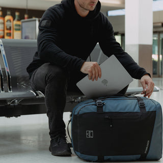 A man sat down with the Tasmin Blue Adventure Bag on the floor, putting a laptop in the bag.