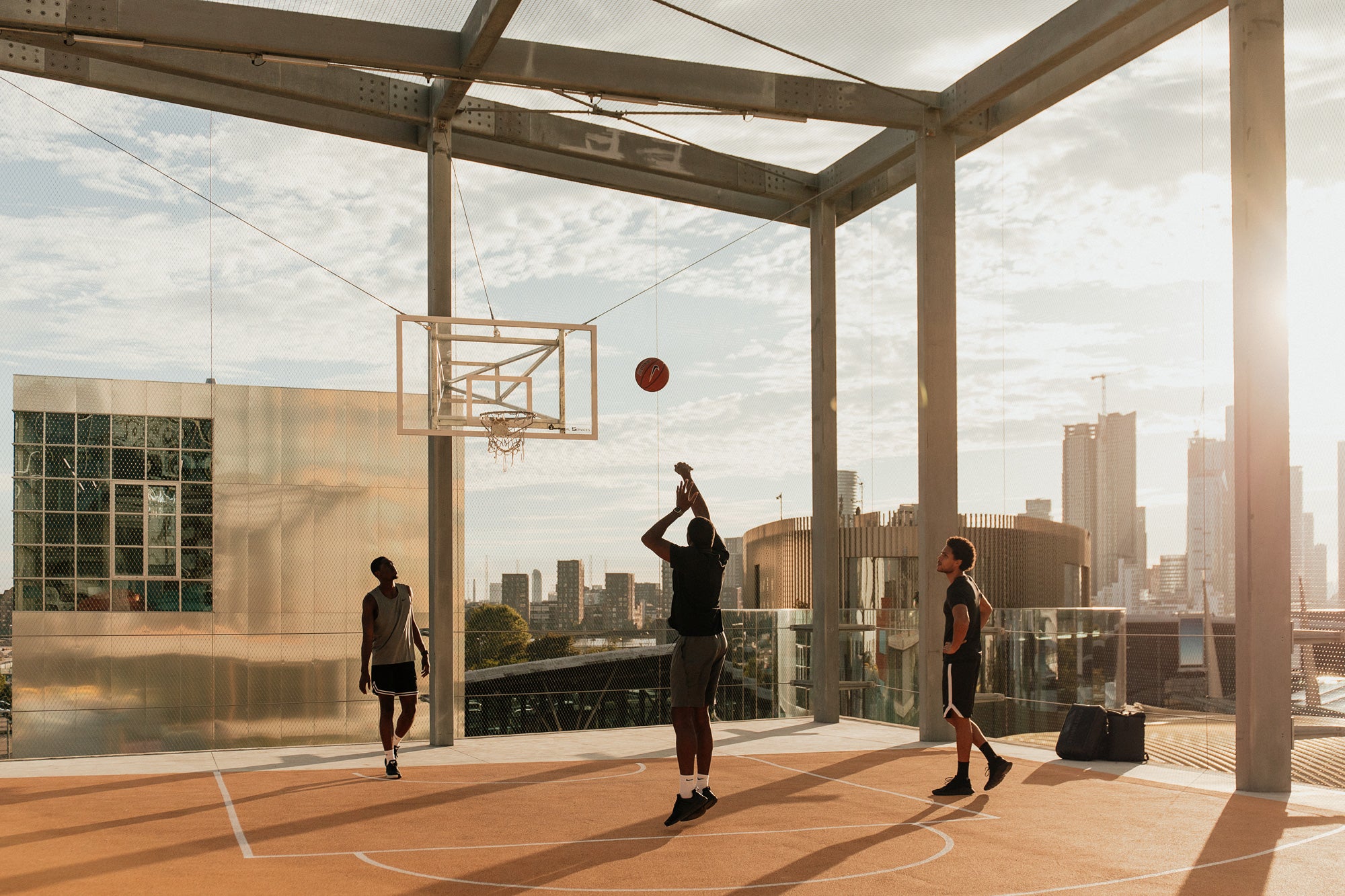 Three people playing basketball on an outdoor court with a city skyline in the background