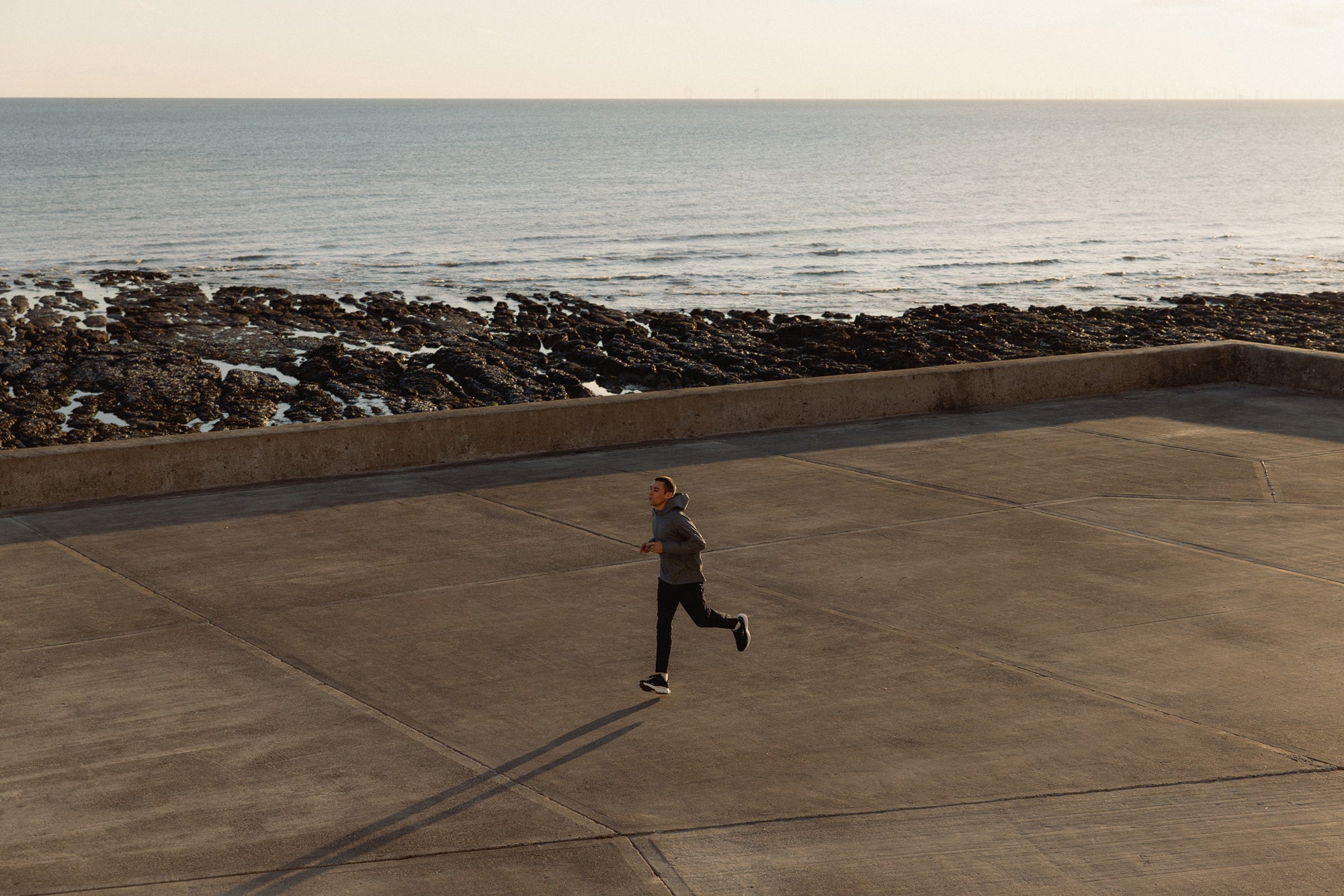 Person running on a concrete path by the ocean with waves crashing against rocks.