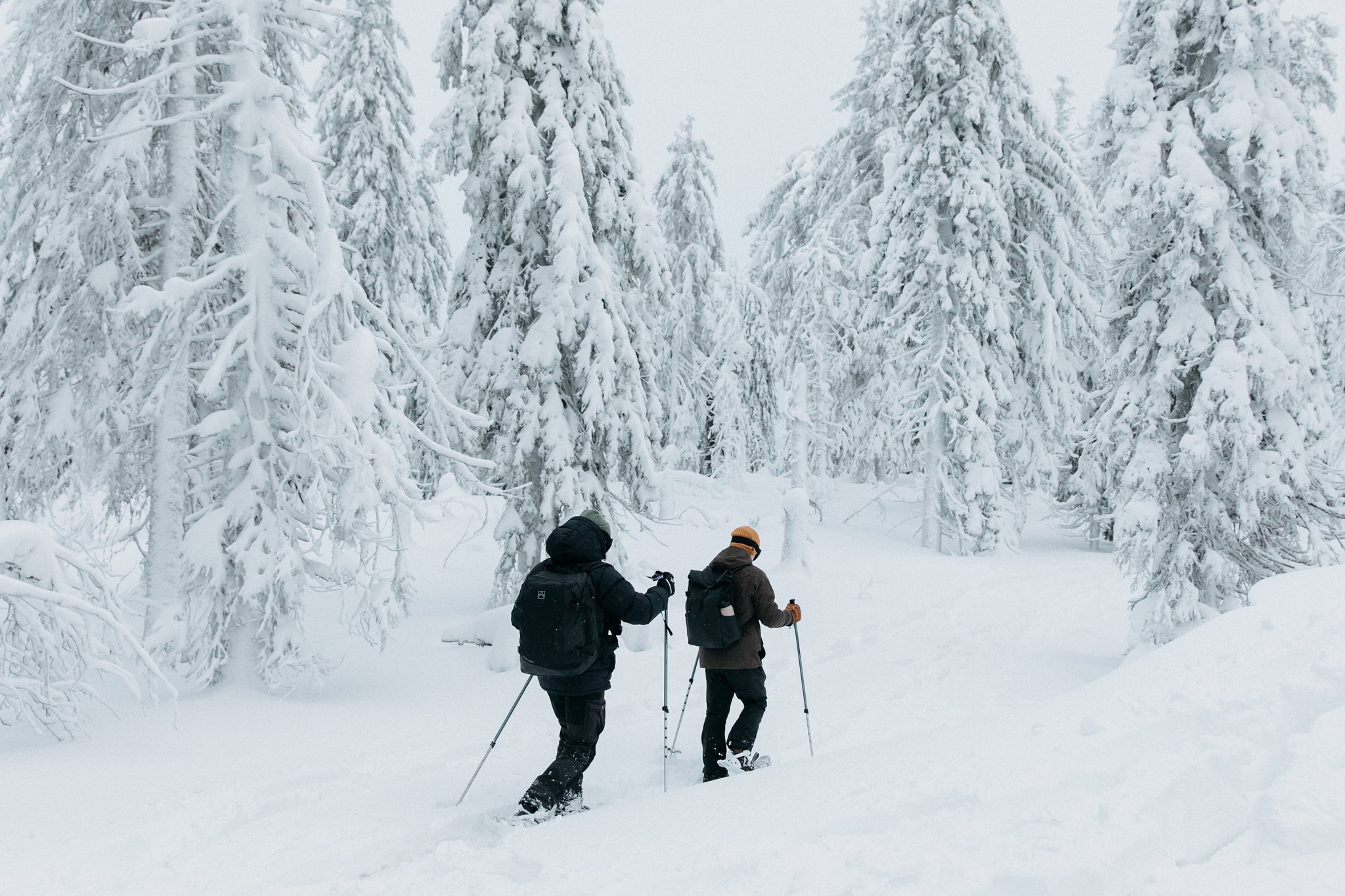 two men walking through the snow wearing skis and black rucksacks and backpacks