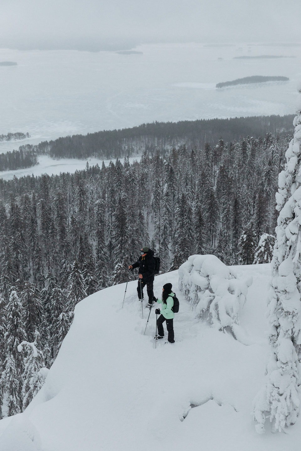 two people at the top of a cliff looking out over a snow covered forest wearing black backpacks and rucksacks