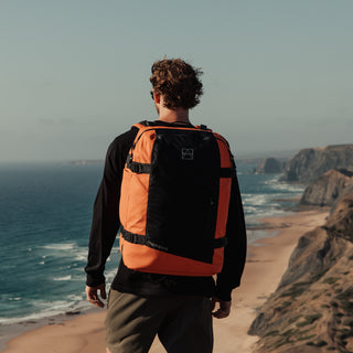 A man standing on a cliff looking out to sea with the Ember Orange Adventure Bag. Shot from the back.