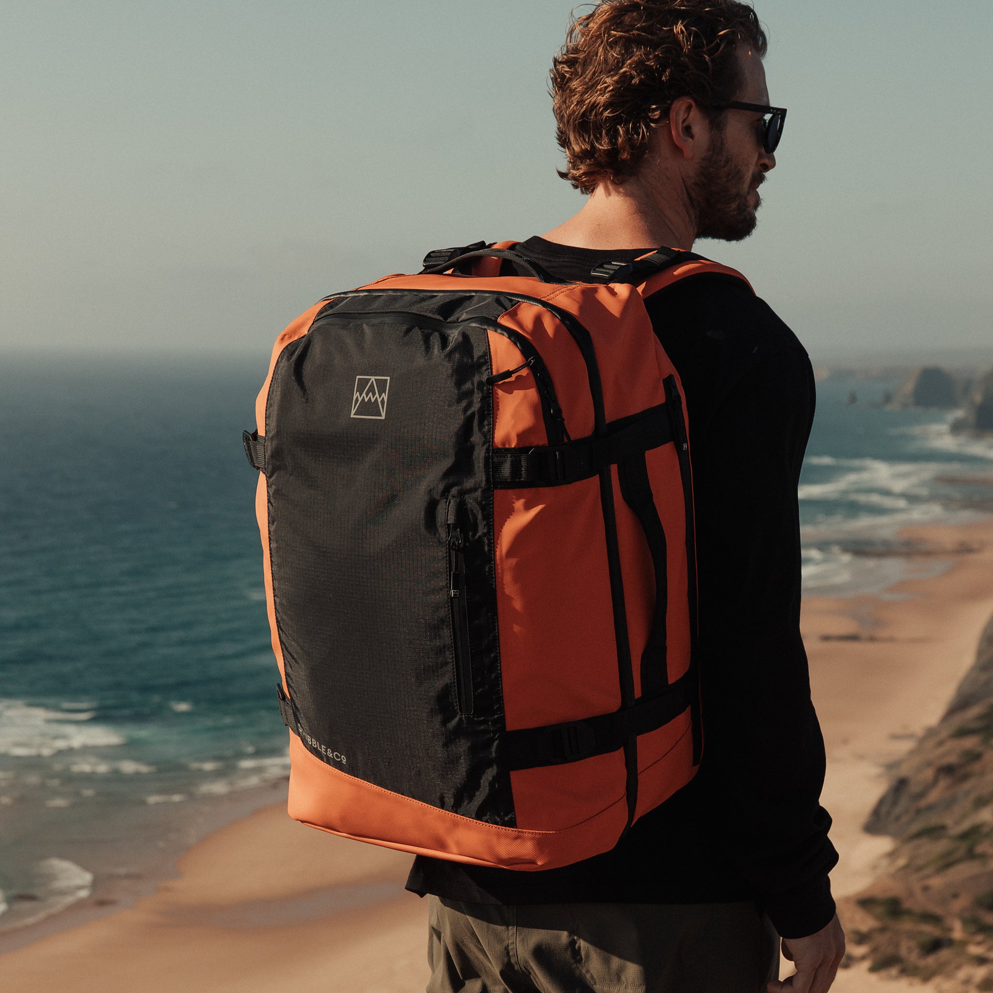 A man standing on a cliff looking out to sea with the Adventure Bag in Ember Orange. Shot from the back.