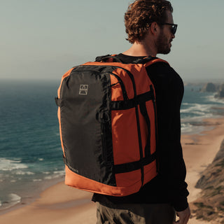 A man standing on a cliff looking out to sea with the Ember Orange Adventure Bag. Shot from the back.