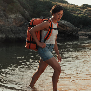 A women walking in the sea up to her ankles. Wearing the Ember Orange Adventure Bag, a white vest top and blue denim shorts.