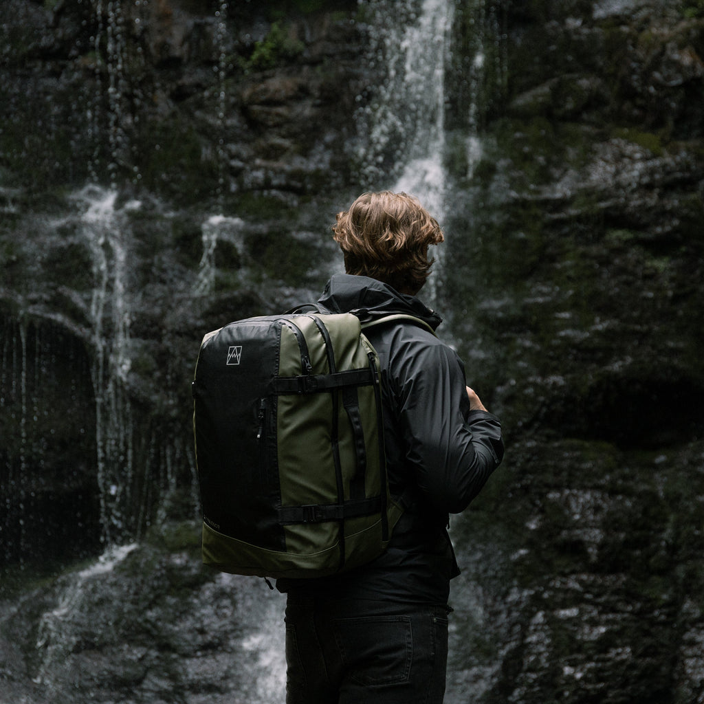 man standing by waterfall wearing a green adventure bag