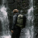 A man wearing the Adventure Bag in Urban Greenwhile looking at a waterfall. Shot from the side.