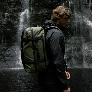 Side view of a man wearing the Urban Green Adventure Bag and a black jacket. Stood in front of a waterfall.