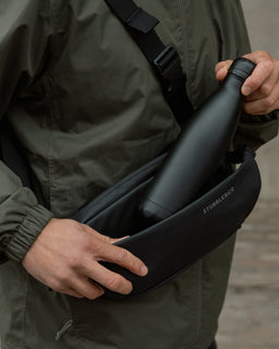 A close up of a man wearing an All Black Crossbody across his chest as he places a black water bottle into the front compartment.