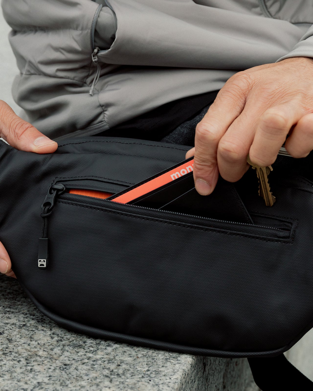 A close up of an All Black Crossbody and someone placing keys and a card holder into the zip pocket on the back of the bag.