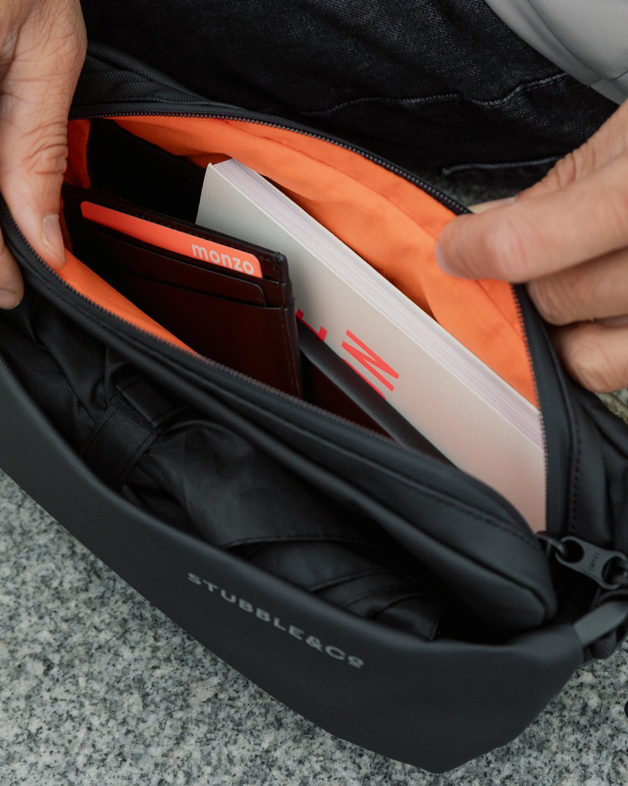 an interior shot of the All Black Crossbody bag showing the orange lining of the bags and the items inside the bag which  are a card holder, a book and an umbrella