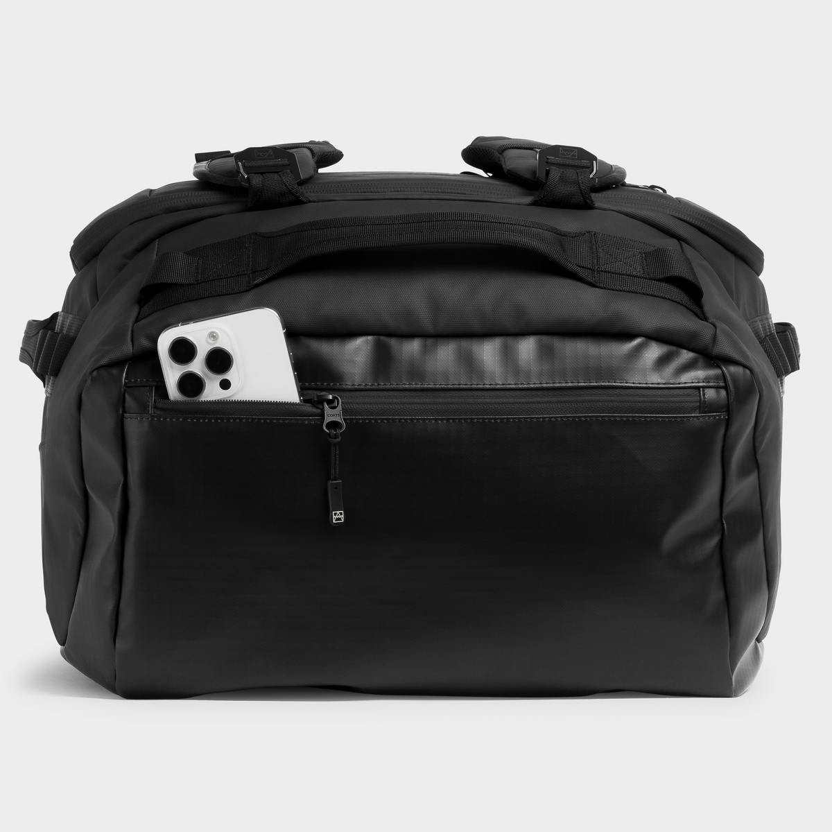 A black nylon duffel bag with a front zippered pocket containing a white smartphone, photographed against a light gray studio background.