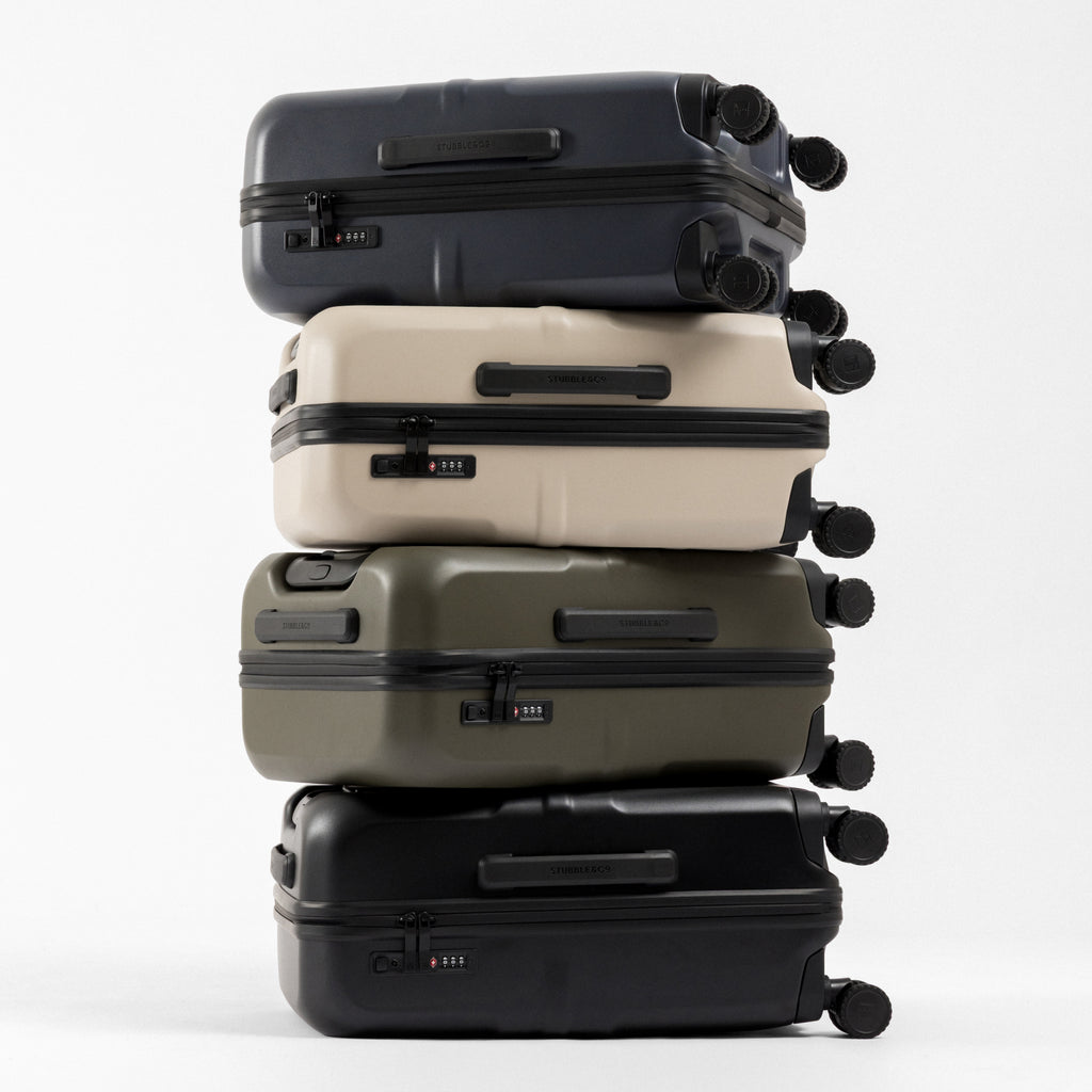 Four hard-shell spinner suitcases stacked vertically on a light gray studio background. From top to bottom, the colors are dark navy, warm cream, olive green, and black.