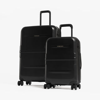 Two black hard-shell spinner suitcases, one larger and one smaller, standing upright on a light gray studio background. [All variants]