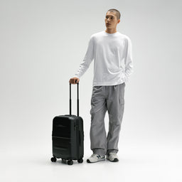 A man wearing a white long-sleeve shirt, gray cargo pants, and white sneakers stands next to a black rolling suitcase, holding its telescoping handle against a light gray studio background. [All variants]