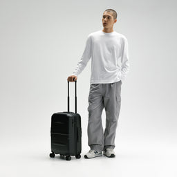 A man wearing a white long-sleeve shirt, gray cargo pants, and white sneakers stands next to a black rolling suitcase, holding its telescoping handle against a light gray studio background. [All variants]