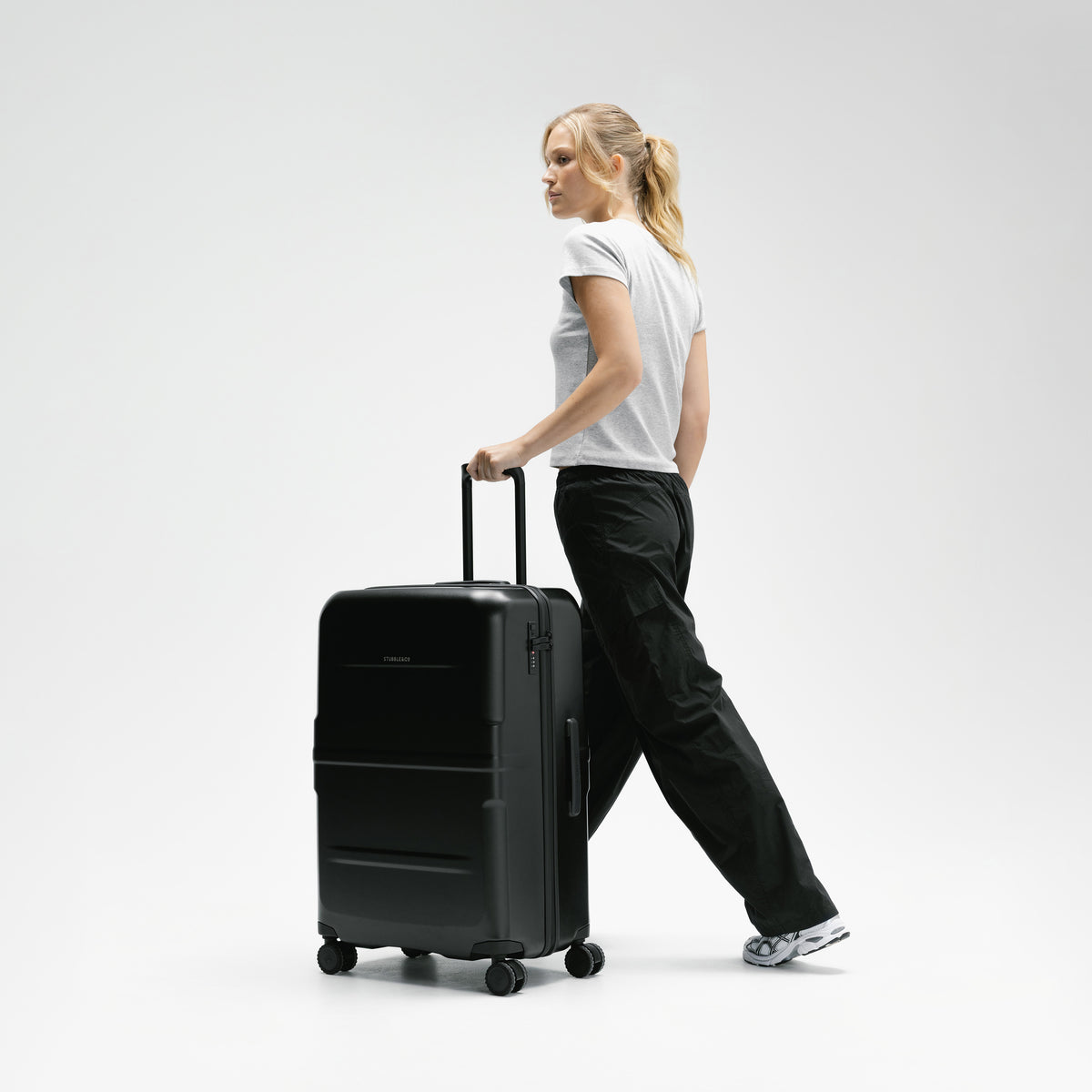 A woman in a white t-shirt and black cargo pants walks while pulling a black hard-shell spinner suitcase against a clean white studio background. [All variants]