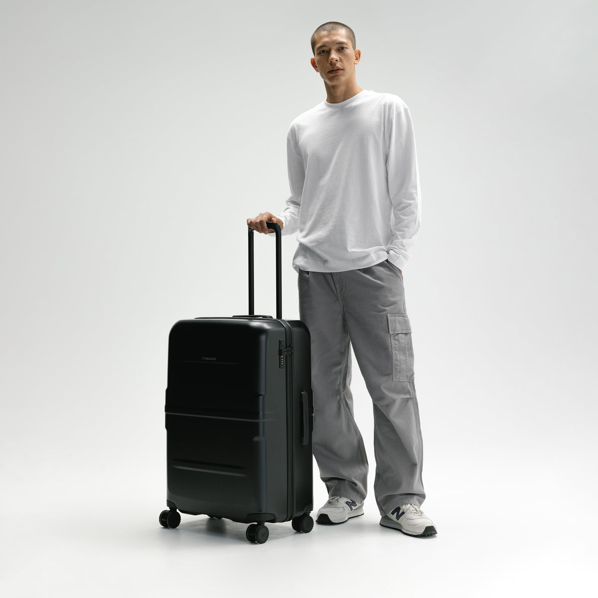A man in a white long-sleeve shirt and gray cargo pants stands next to a matte black hard-shell spinner suitcase, holding its telescoping handle in a studio setting with a light gray background. [All variants]