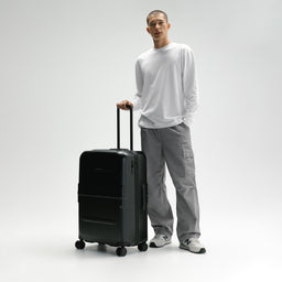 A man in a white long-sleeve shirt and gray cargo pants stands next to a matte black hard-shell spinner suitcase, holding its telescoping handle in a studio setting with a light gray background. [All variants]