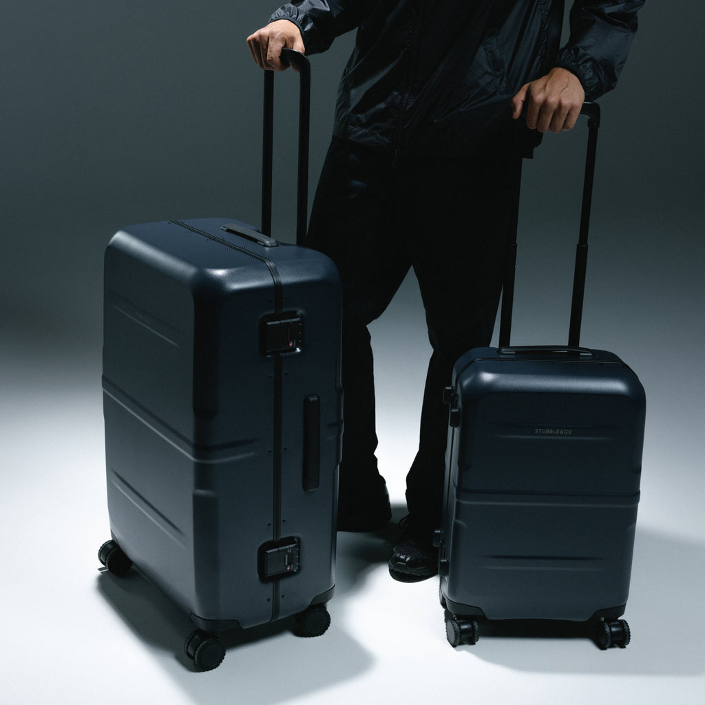Two matte black hard-shell rolling suitcases, one large and one small, shown with a person in a black jacket and pants standing behind them against a dark studio background.
