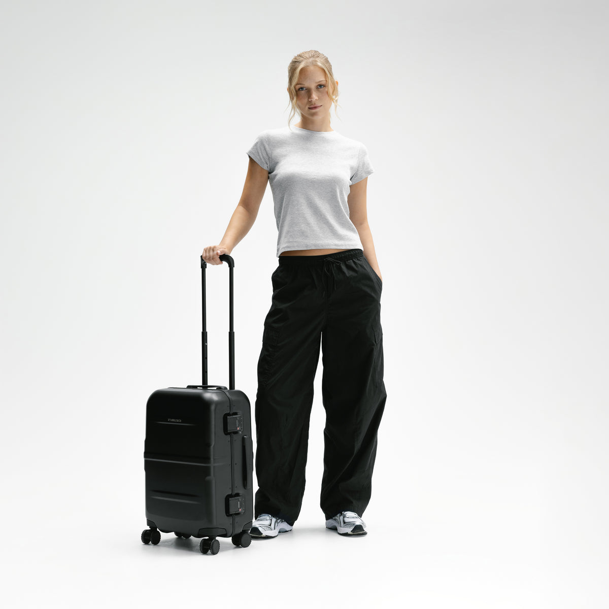 A woman stands in a studio wearing a white cropped T-shirt, loose black pants, and sneakers, holding the handle of a black hard-shell rolling suitcase against a light gray background. [All variants]