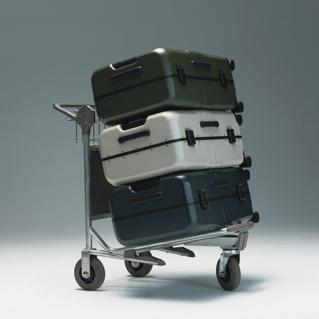 Three hard-shell suitcases stacked on a metal luggage cart against a smooth gray studio background.